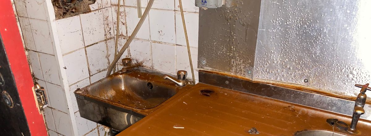 A derelict commercial kitchen area featuring a stained stainless steel sink and countertop against a white tiled wall heavily affected by damp, mold, and debris.