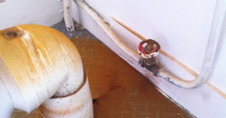 A rusty valve attached to a white pipe runs along a wall, with another large, white, L-shaped pipe nearby. The floor beneath has brown stains and patches showing signs of dry rot spore dust