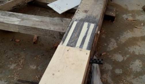 A long timber beam with a light-colored new section spliced onto an older, weathered beam; the joint has three visible dark adhesive or reinforcement strips.
