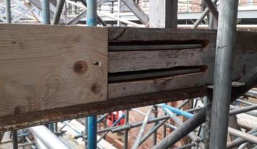 Close-up of a wooden beam prepared for timber resin splice installation, with several slots cut into it. Metal scaffolding surrounds the beam at a construction site, with brick walls and more scaffolding in the background.